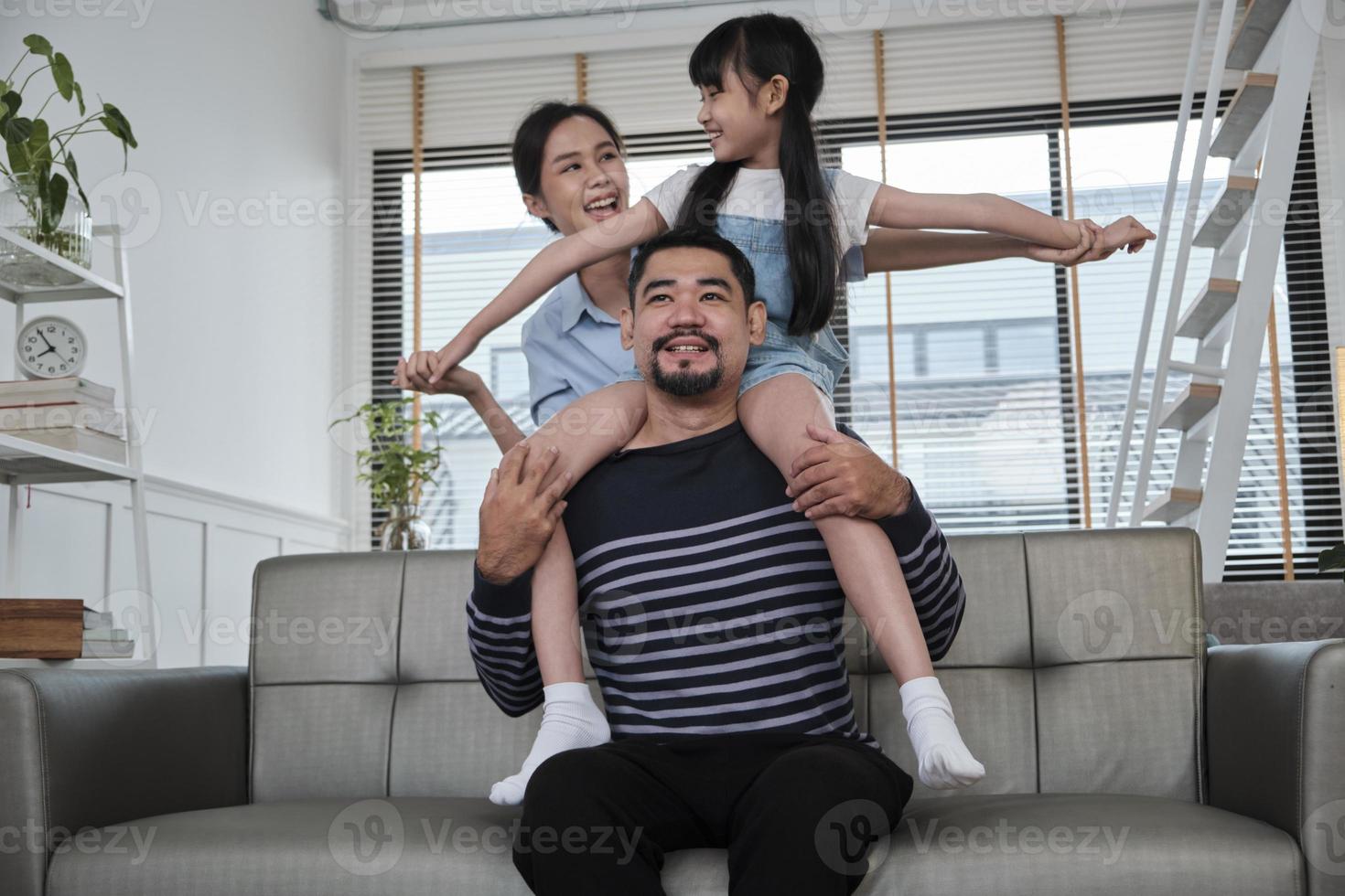Asian Thai family together, dad plays and teases with daughter and mum by carrying and holding girl on shoulders in home living room, happy leisure times, lovely weekend, wellbeing domestic lifestyle. photo