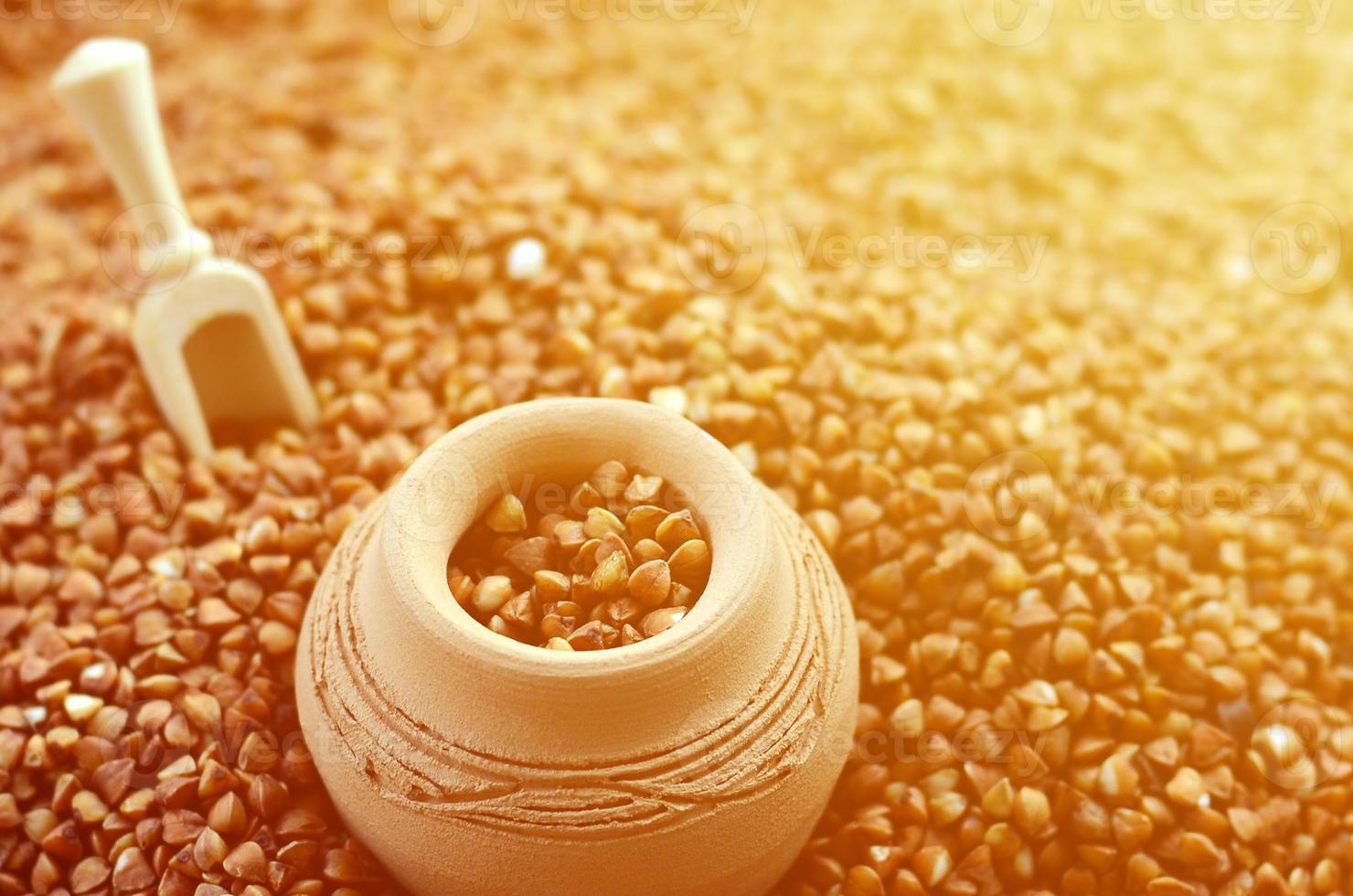 Background image of a large pile of buckwheat, in the middle of which lies a small jug and a wooden spatula for cereals photo