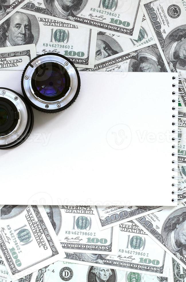 dos lentes fotográficos y un cuaderno blanco se encuentran en el fondo de muchos billetes de dólar. espacio para texto foto