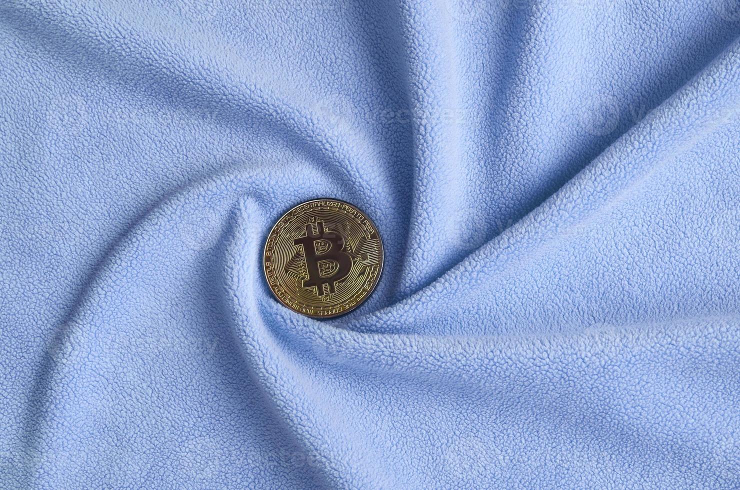 The golden bitcoin lies on a blanket made of soft and fluffy light blue fleece fabric with a large number of relief folds. The shape of the folds resembles a fan from a video card cooler photo