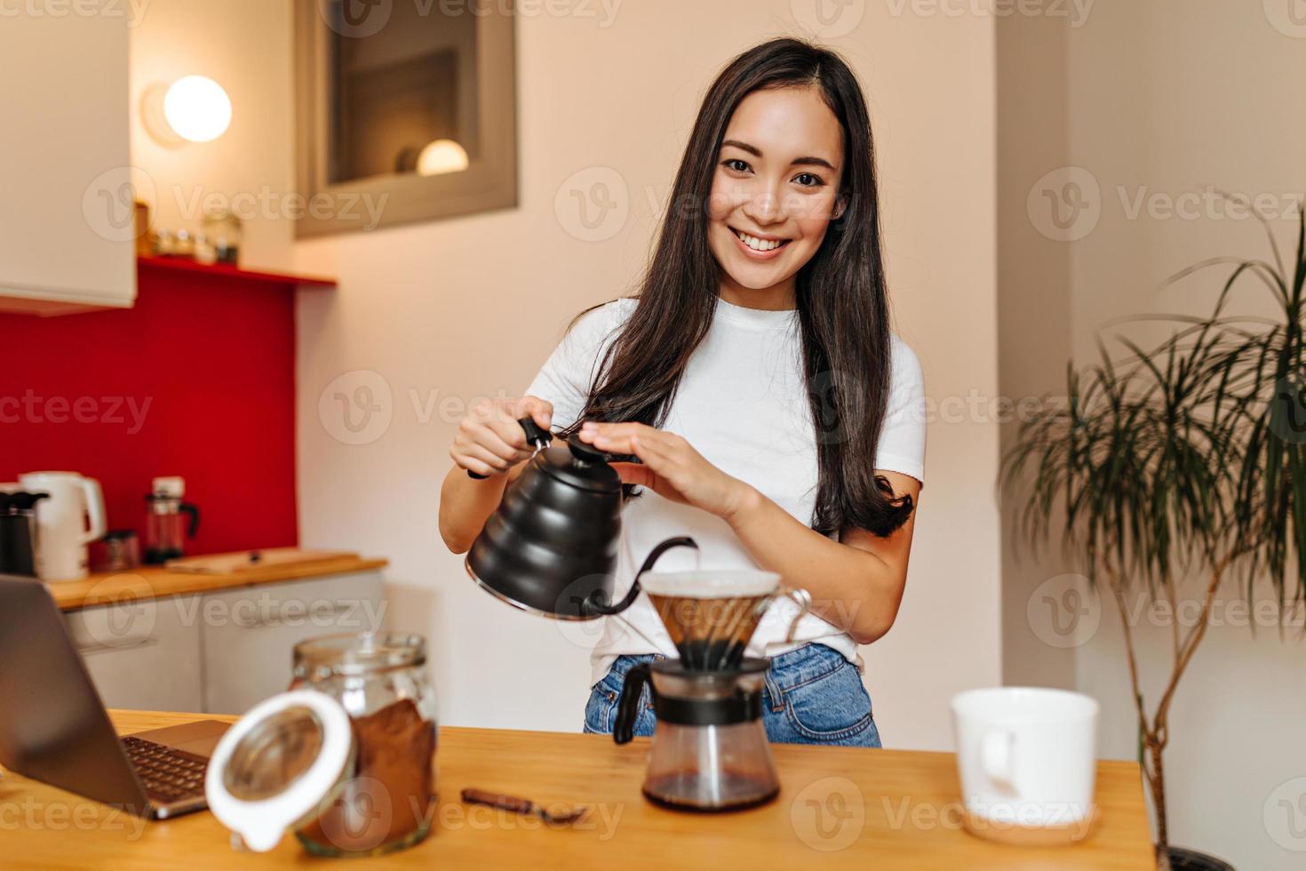chica de ojos marrones con camiseta blanca mira a la cámara con una sonrisa y vierte agua hirviendo en una cafetera foto