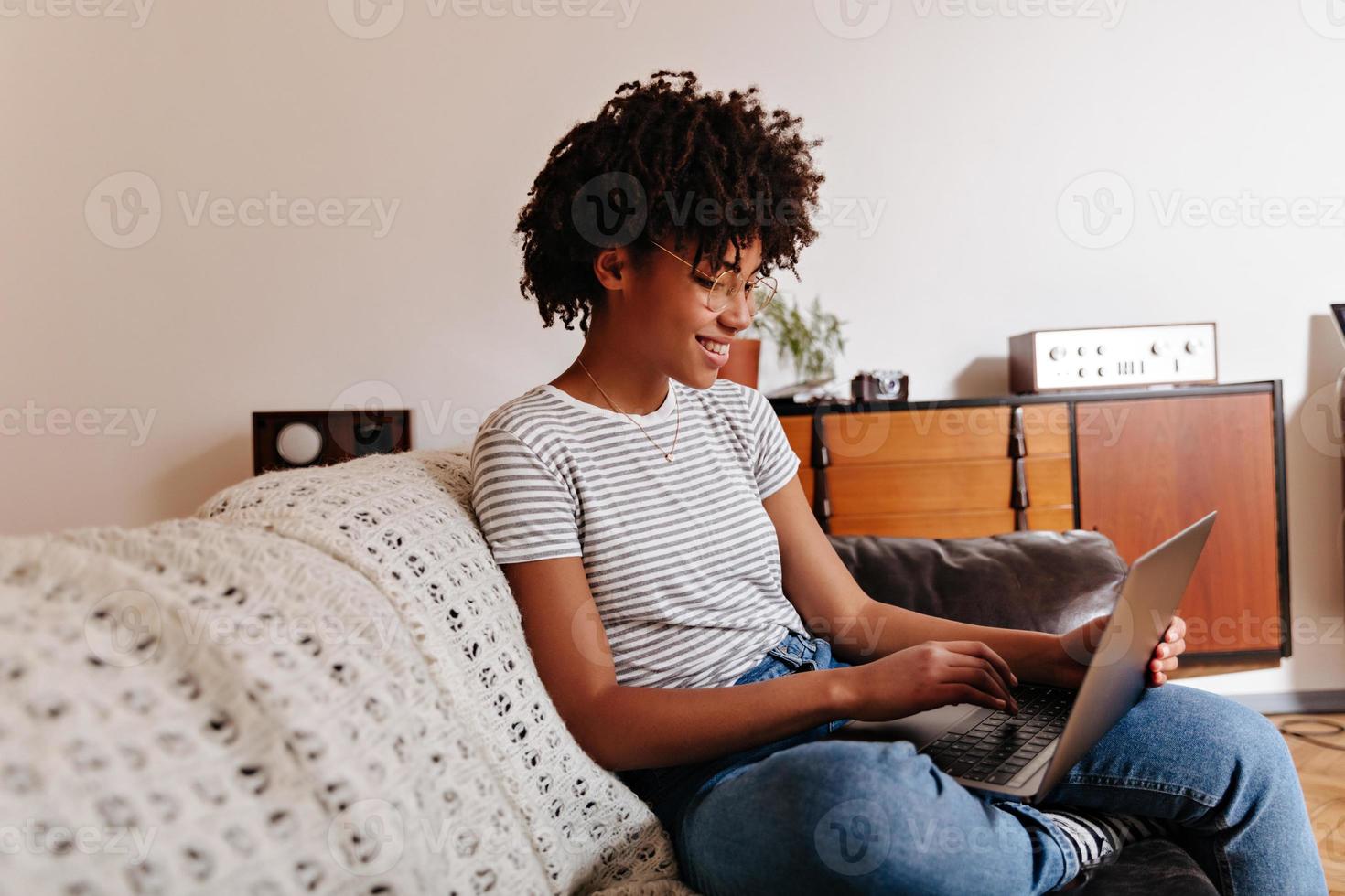 una chica africana con una camiseta a rayas está sentada en el sofá y charlando con una sonrisa en una laptop. foto