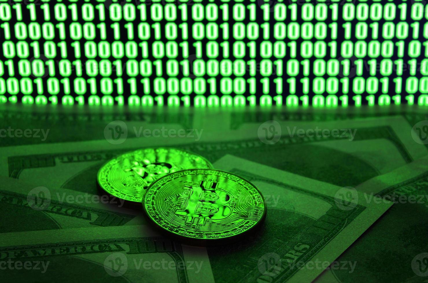 Two bitcoins lies on a pile of dollar bills on the background of a monitor depicting a binary code of bright green zeros and one units on a black background. Low key lighting photo