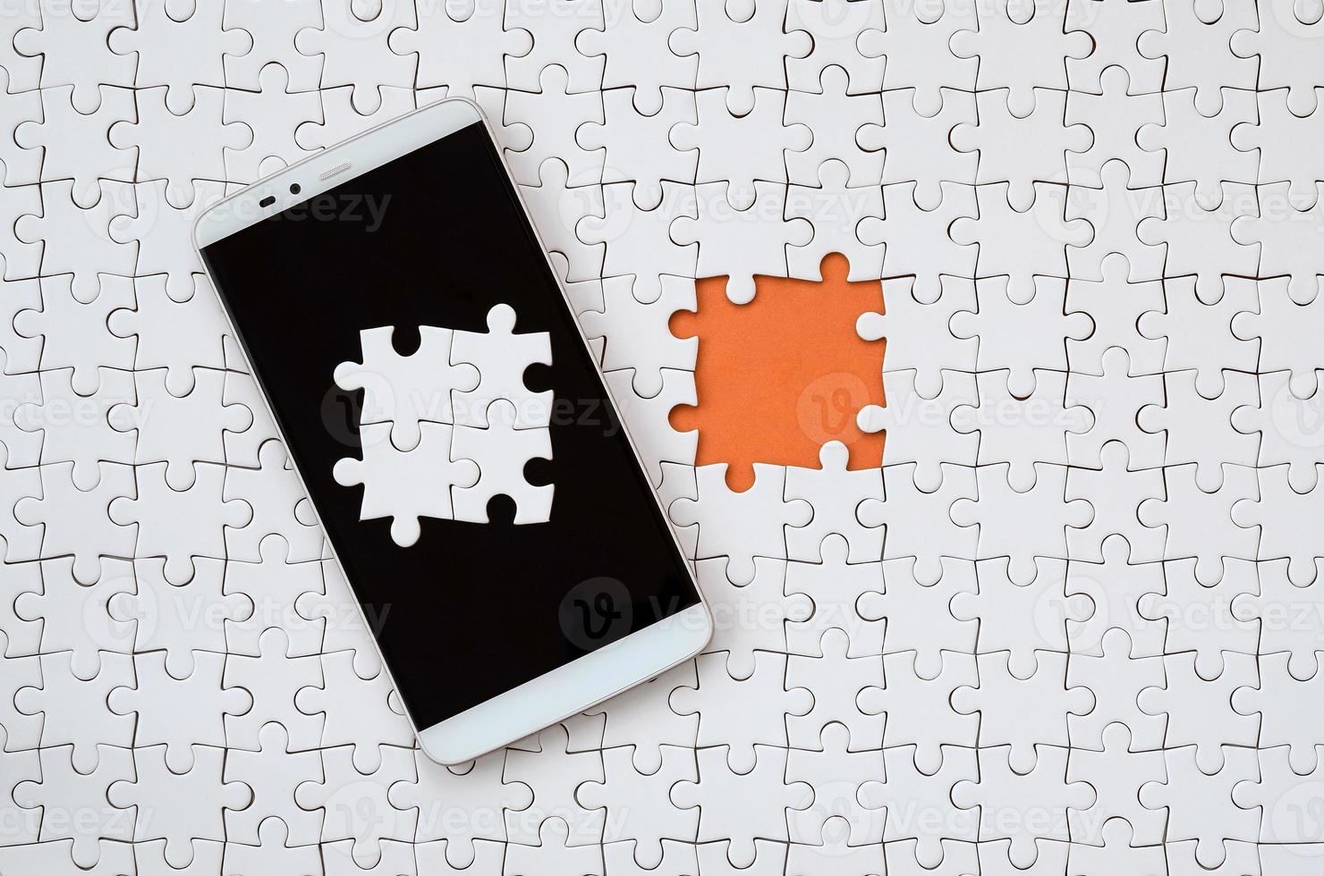 A modern big smartphone with several puzzle elements on the touch screen lies on a white jigsaw puzzle in an assembled state with missing elements photo