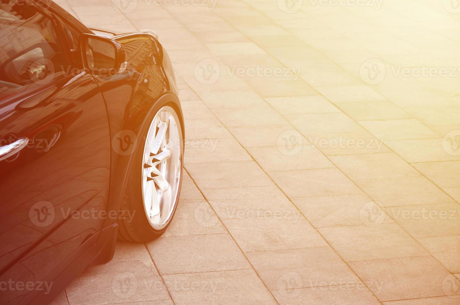 Diagonal view of a black glossy car with white wheels, which stands on a square of gray tiles photo