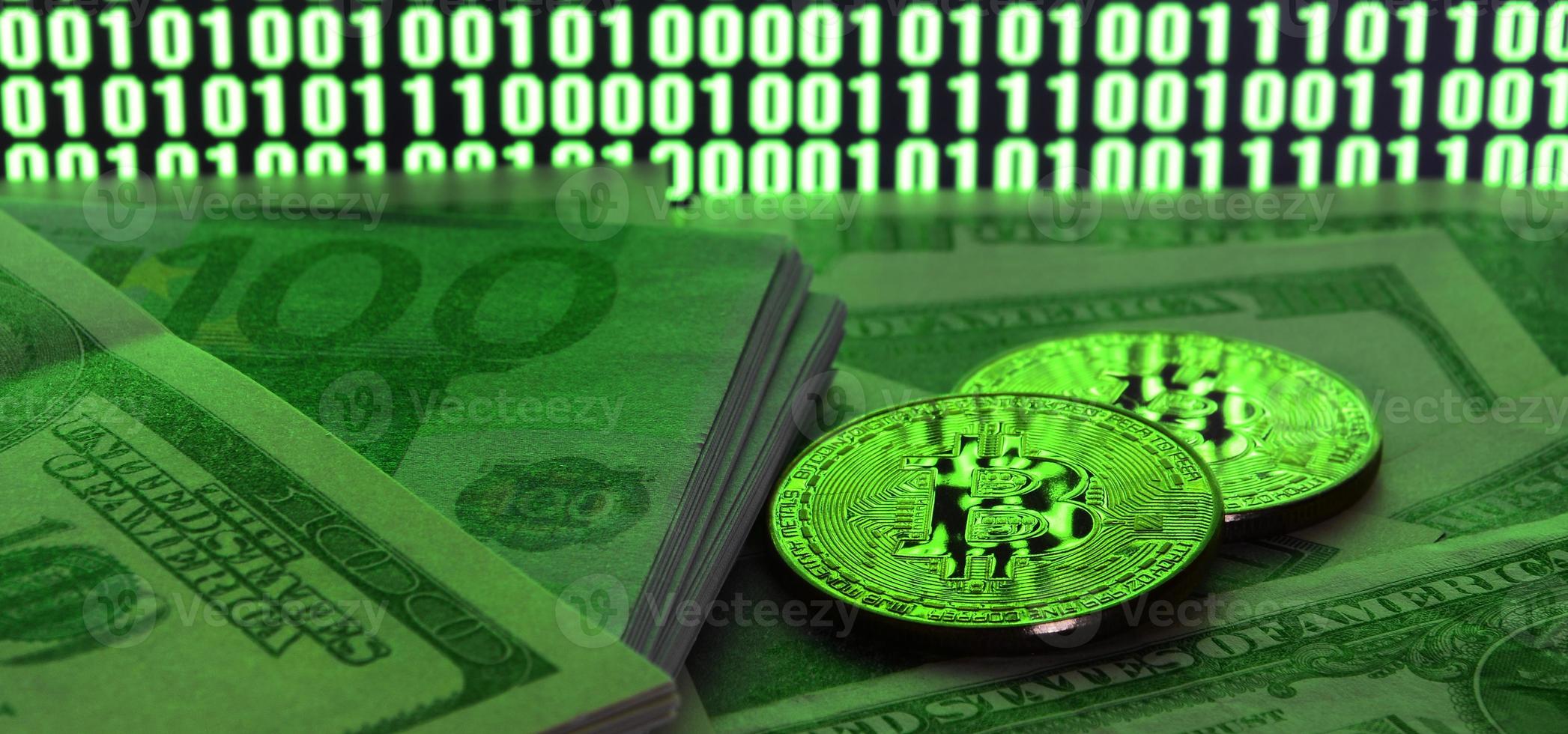 Two bitcoins lies on a pile of dollar bills on the background of a monitor depicting a binary code of bright green zeros and one units on a black background. Low key lighting photo