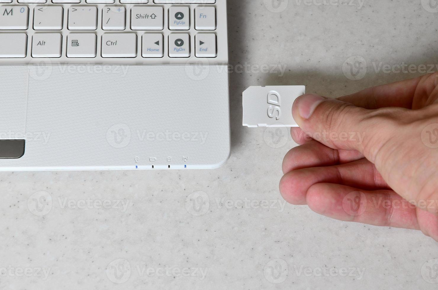 A male hand inserts a white compact SD card into the corresponding input in the side of the white netbook. Man uses modern technologies to store memory and digital data photo