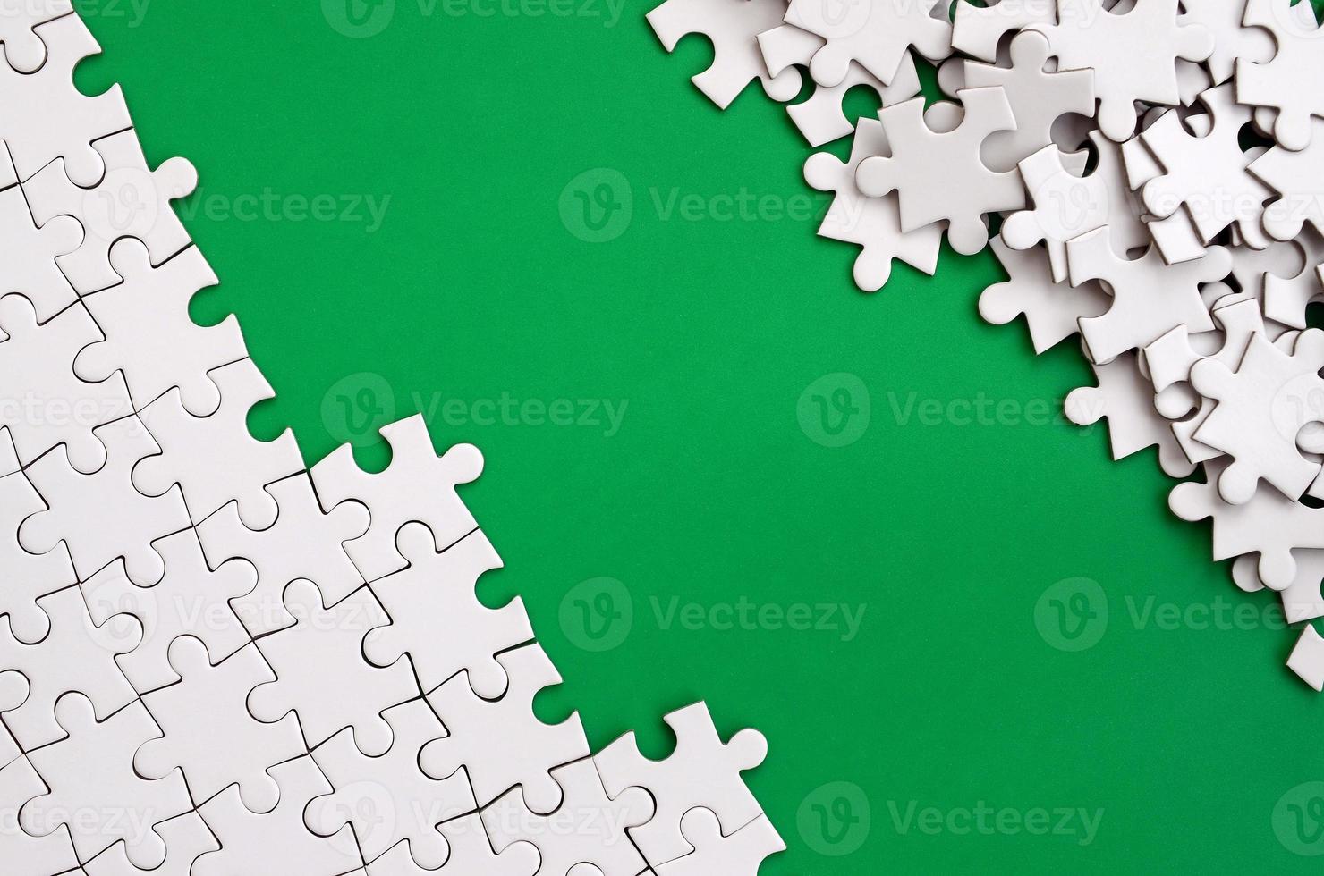 Fragment of a folded white jigsaw puzzle and a pile of uncombed puzzle elements against the background of a green surface. Texture photo with space for text