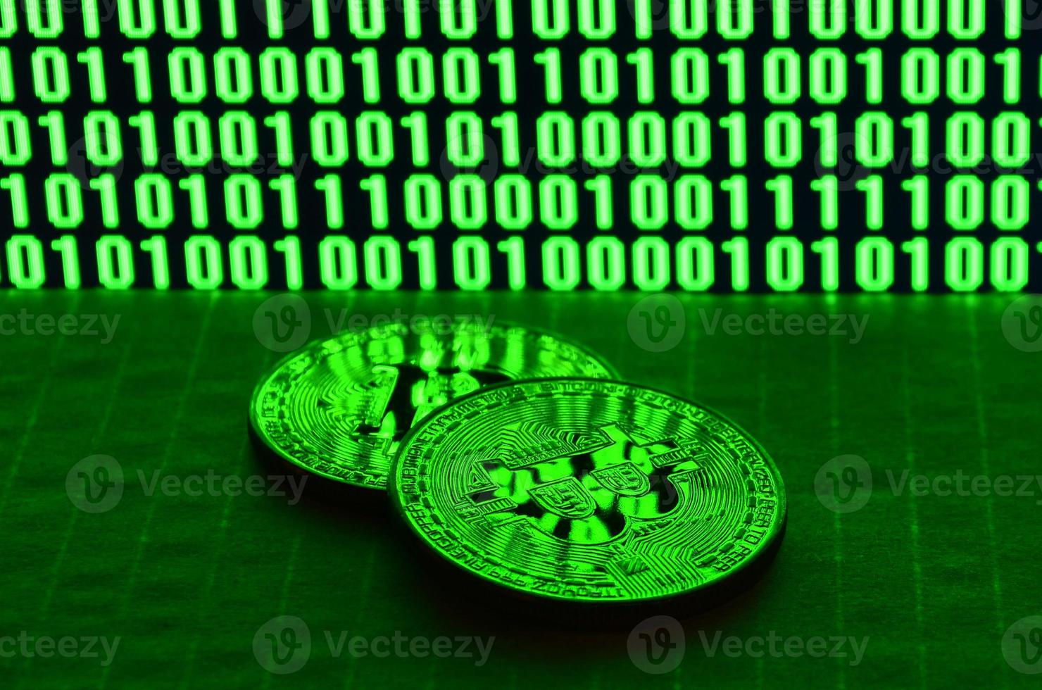 A pair of bitcoins lies on a cardboard surface on the background of a monitor depicting a binary code of bright green zeros and one units on a black background. Low key lighting photo