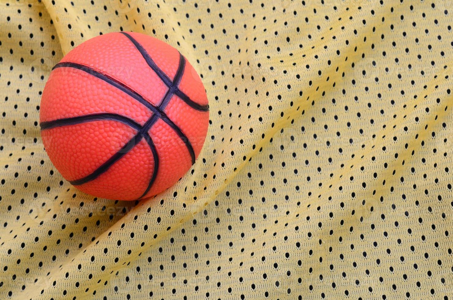 Small orange rubber basketball lies on a yellow sport jersey clothing fabric texture and background with many folds photo