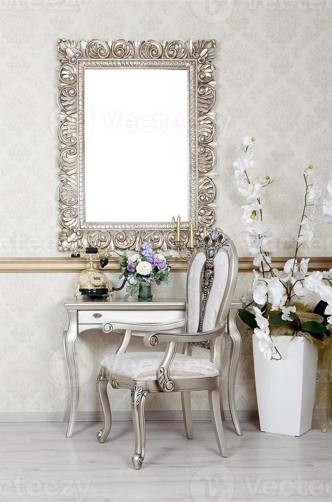 A fragment of a retro interior with a chair and table, on which is a telephone and a vase of flowers. Above the table hangs an empty picture frame for your photo