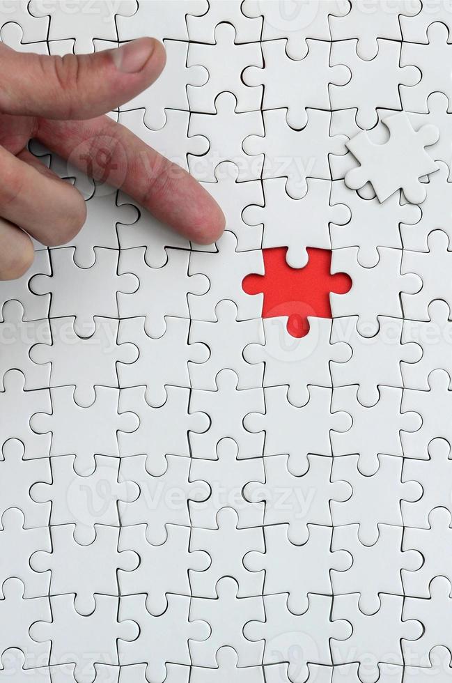 The texture of a white jigsaw puzzle in the assembled state with one missing element, forming a red space, pointed to by the finger of the male hand photo