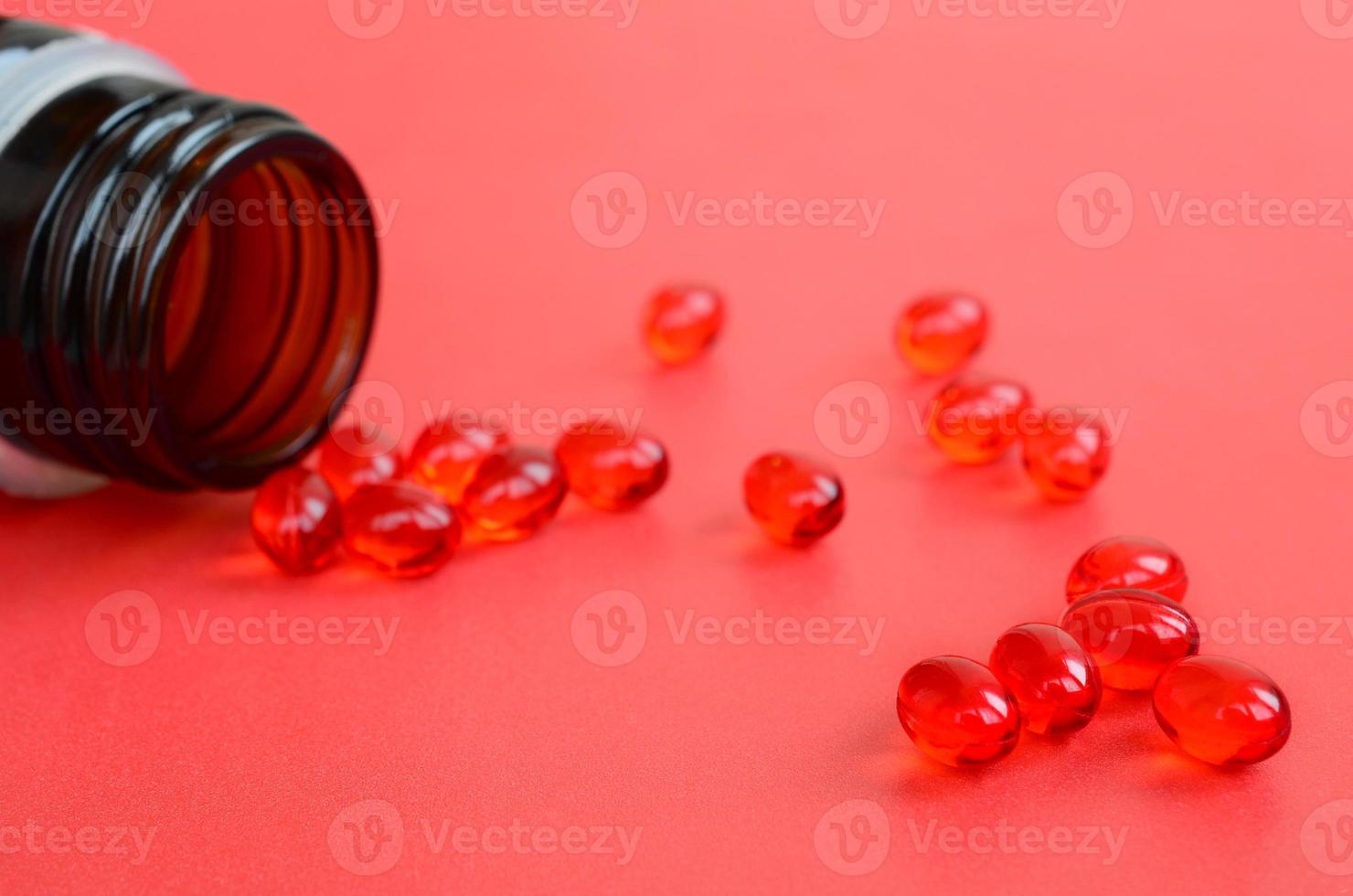 A lot of transparent red tablets were scattered from a small glass brown jar on a red surface photo