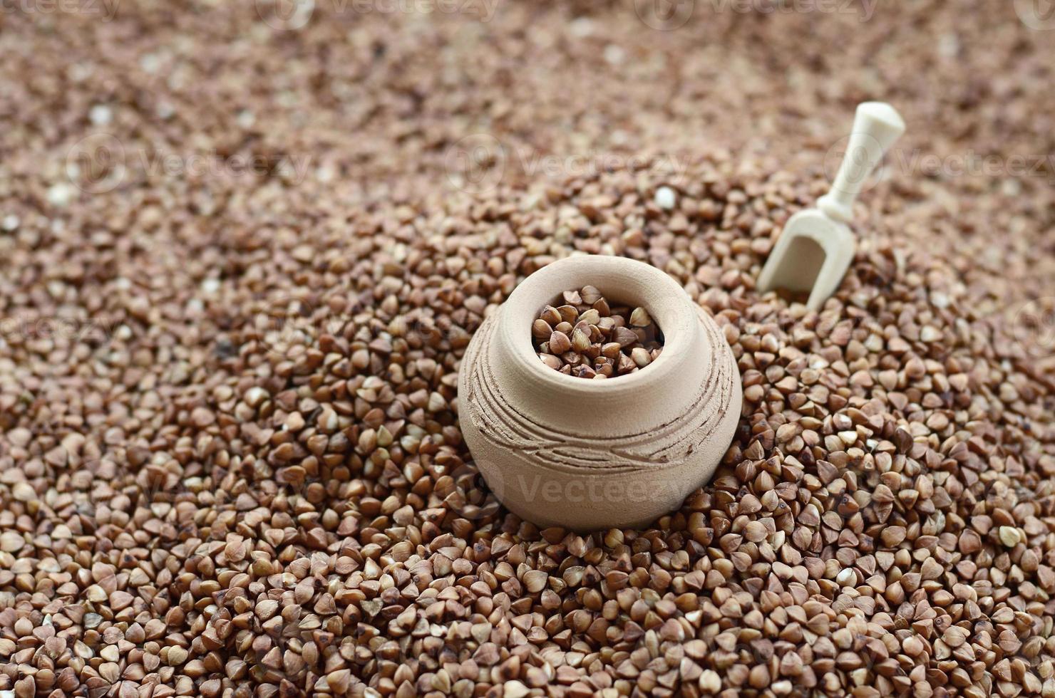 Background image of a large pile of buckwheat, in the middle of which lies a small jug and a wooden spatula for cereals photo