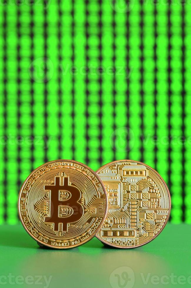 Two gold bitcoins lie on the green surface on the background of the display, which shows the process of mining the crypto currency photo