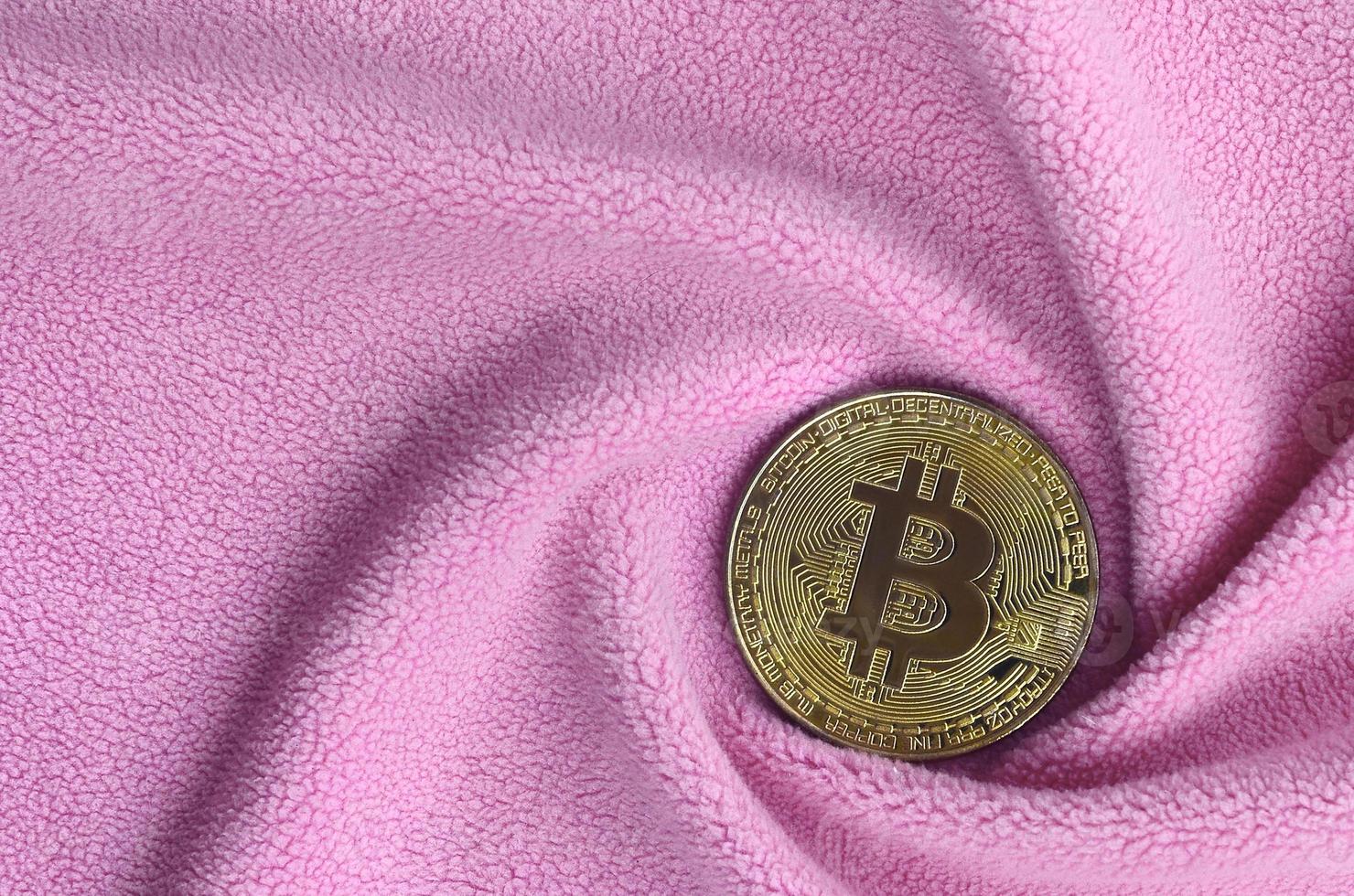 The golden bitcoin lies on a blanket made of soft and fluffy light pink fleece fabric with a large number of relief folds. The shape of the folds resembles a fan from a video card cooler photo