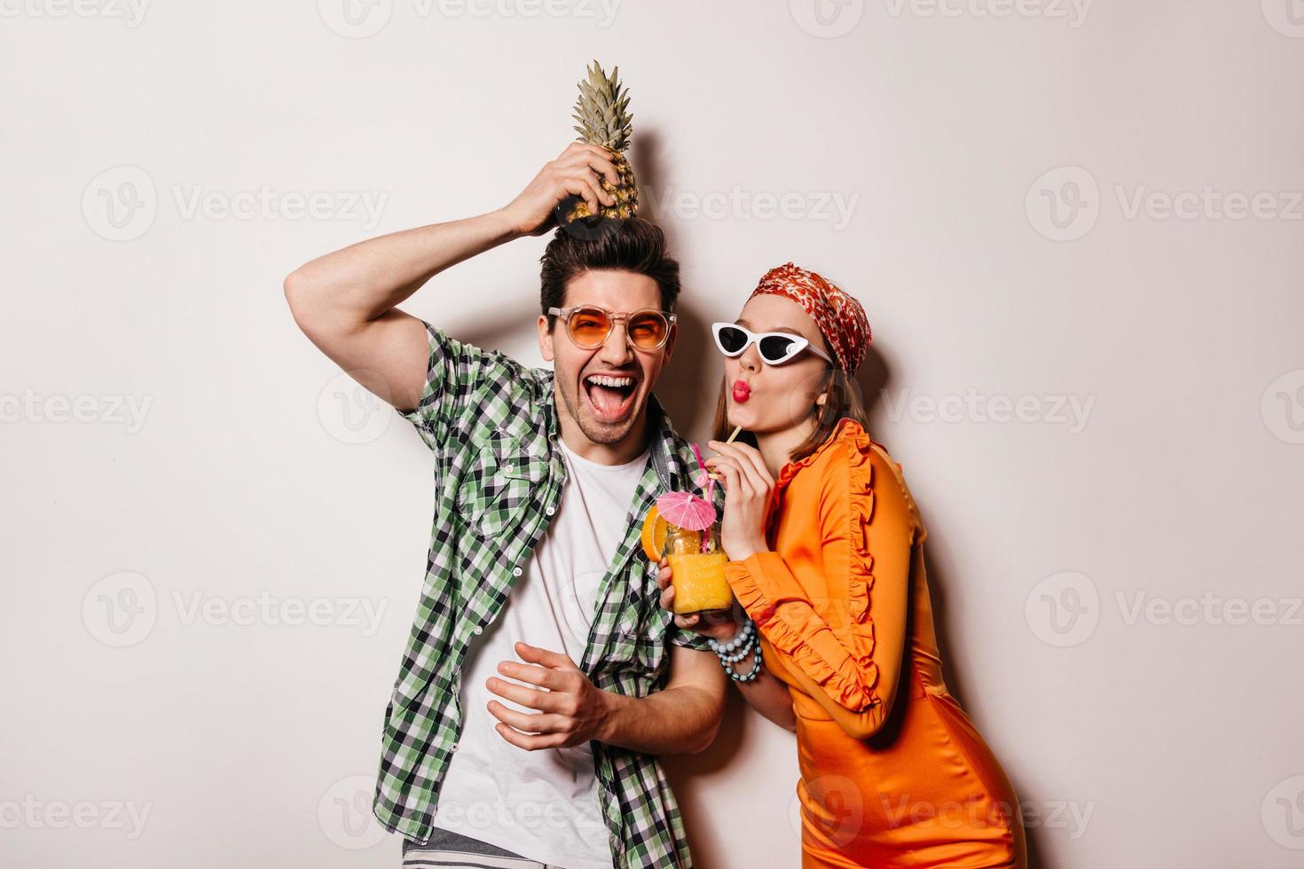 Man in orange glasses holds pineapple on his head and laughs. Woman in bright dress and sunglasses photo