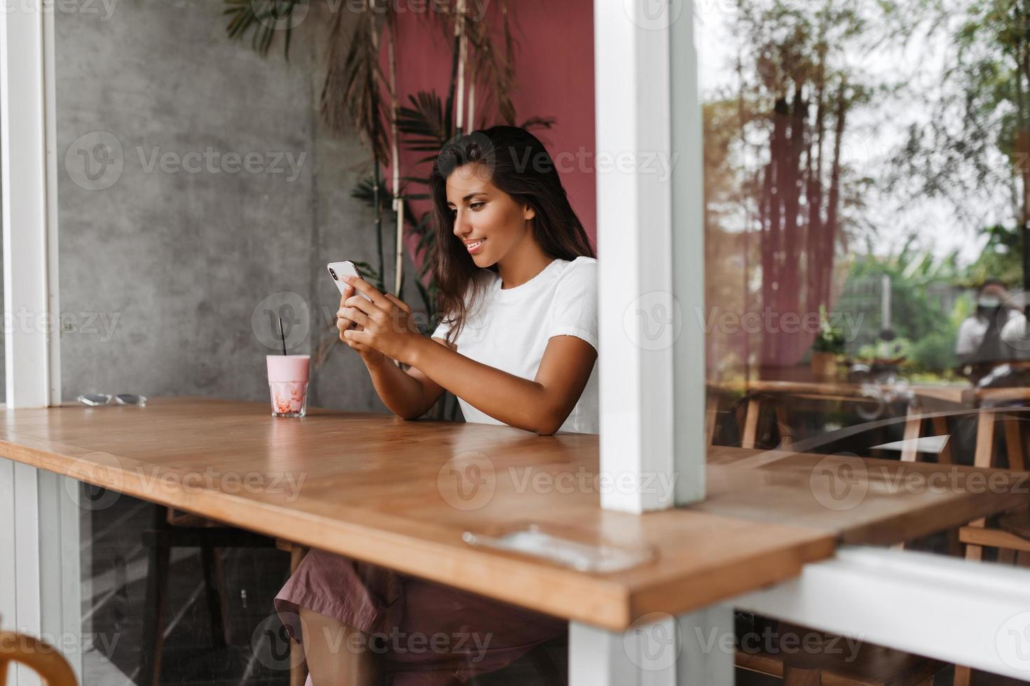 la mujer se sienta a la mesa en un acogedor café y charla en un teléfono inteligente. retrato de chica bronceada con camiseta blanca foto