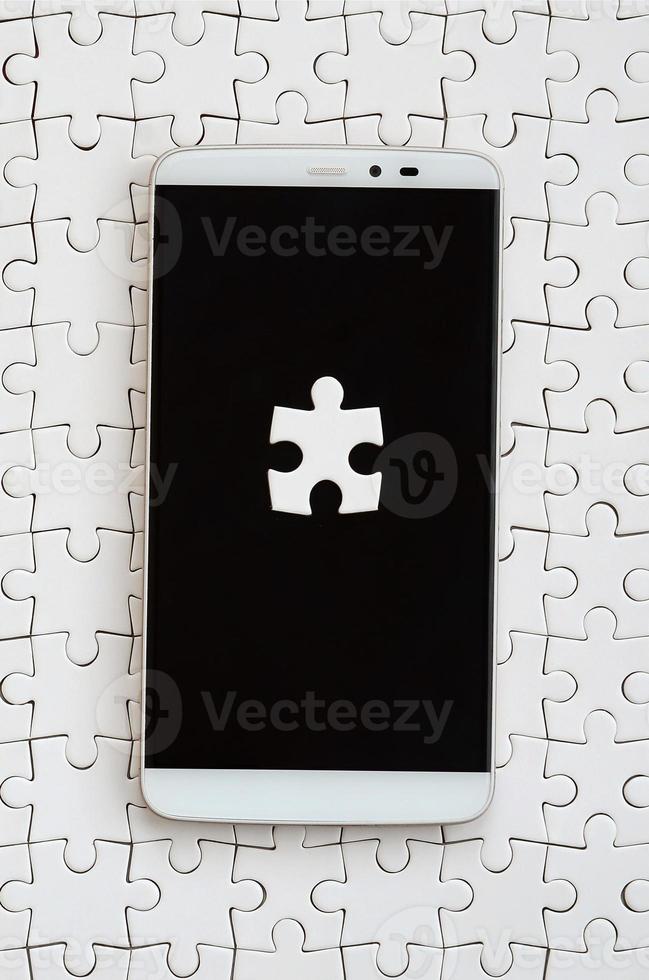 A modern big smartphone with several puzzle elements on the touch screen lies on a white jigsaw puzzle in an assembled state photo