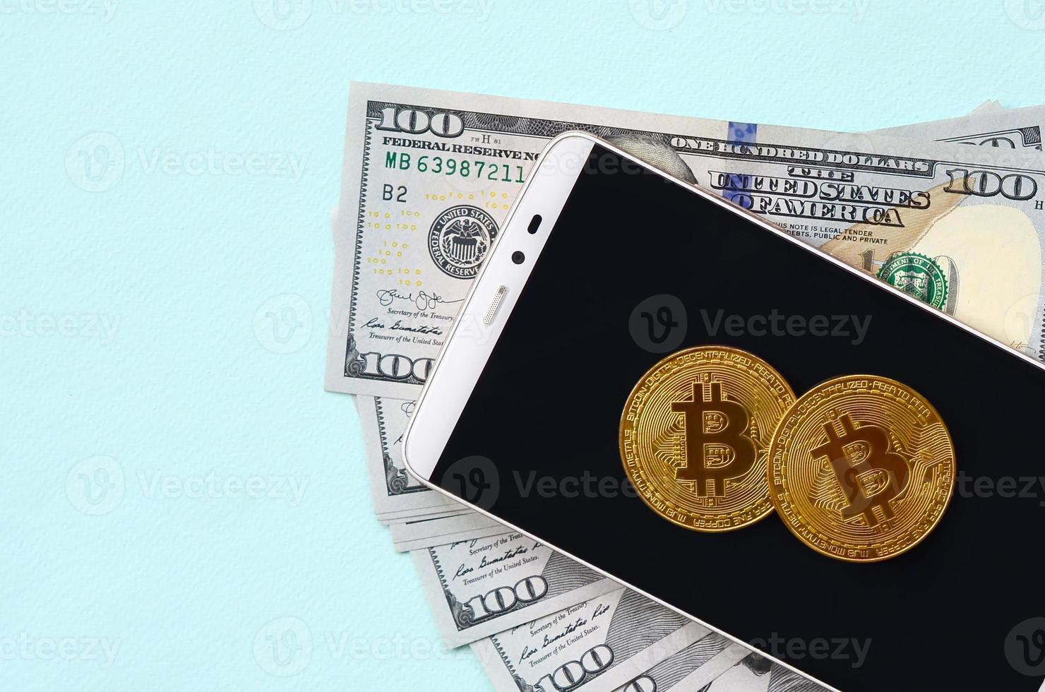 Bitcoins lies on a smartphone and hundred dollar bills on a light blue background photo