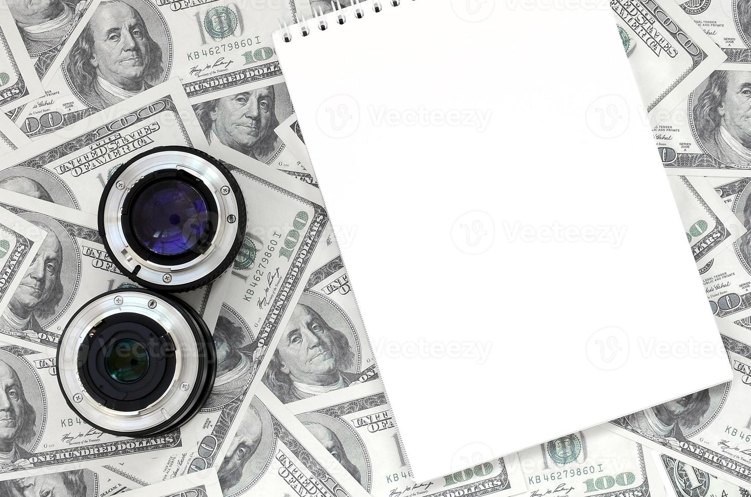 Two photographic lenses and white notebook lie on the background of a lot of dollar bills. Space for text photo