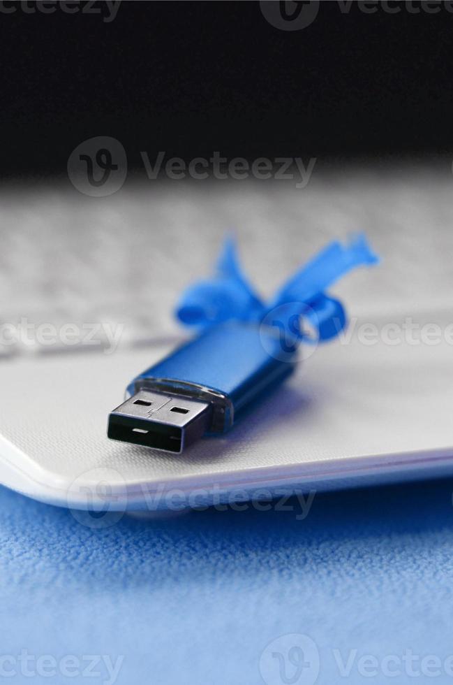 Brilliant blue usb flash memory card with a blue bow lies on a blanket of soft and furry light blue fleece fabric beside to a white laptop. Classic female gift design for a memory card photo