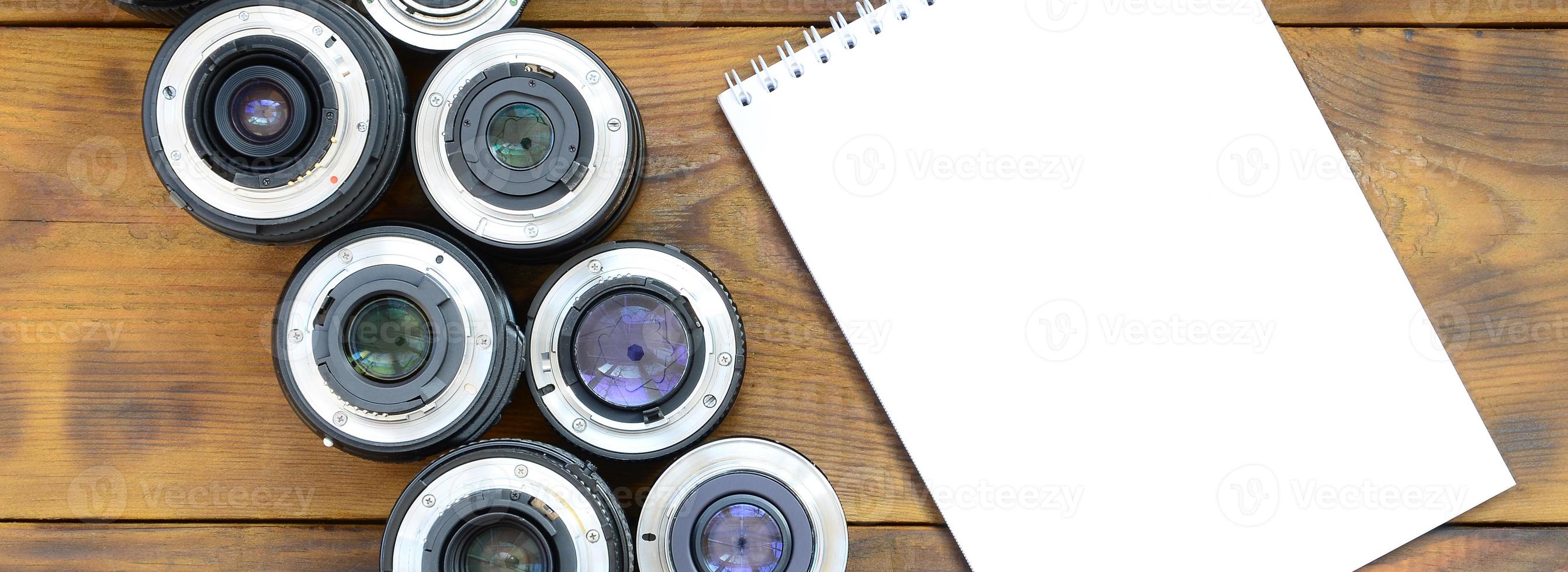 Several photographic lenses and white notebook lie on a brown wooden background. Space for text photo