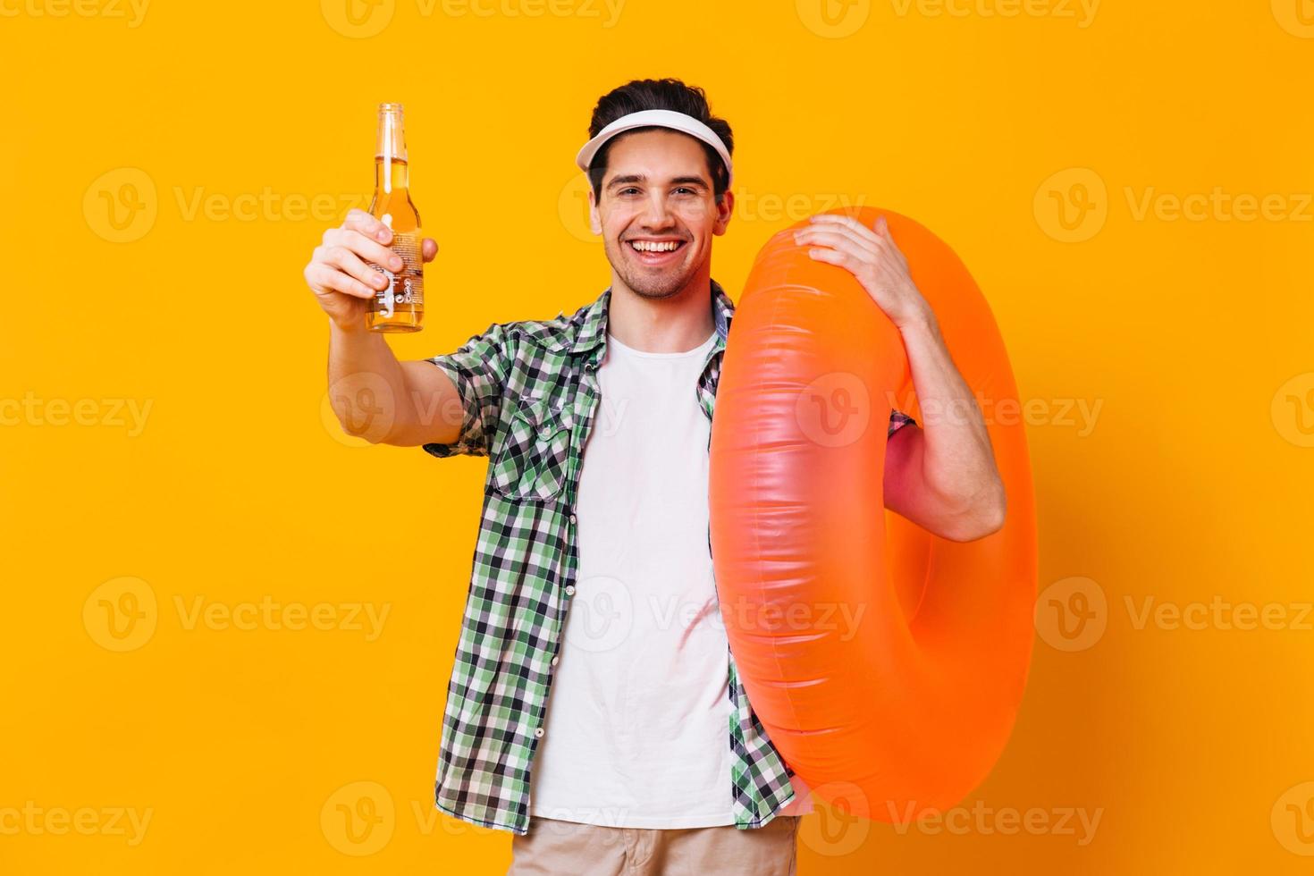 Portrait of man in cap, shirt and t-shirt holding bottle of beer and inflatable circle on isolated photo