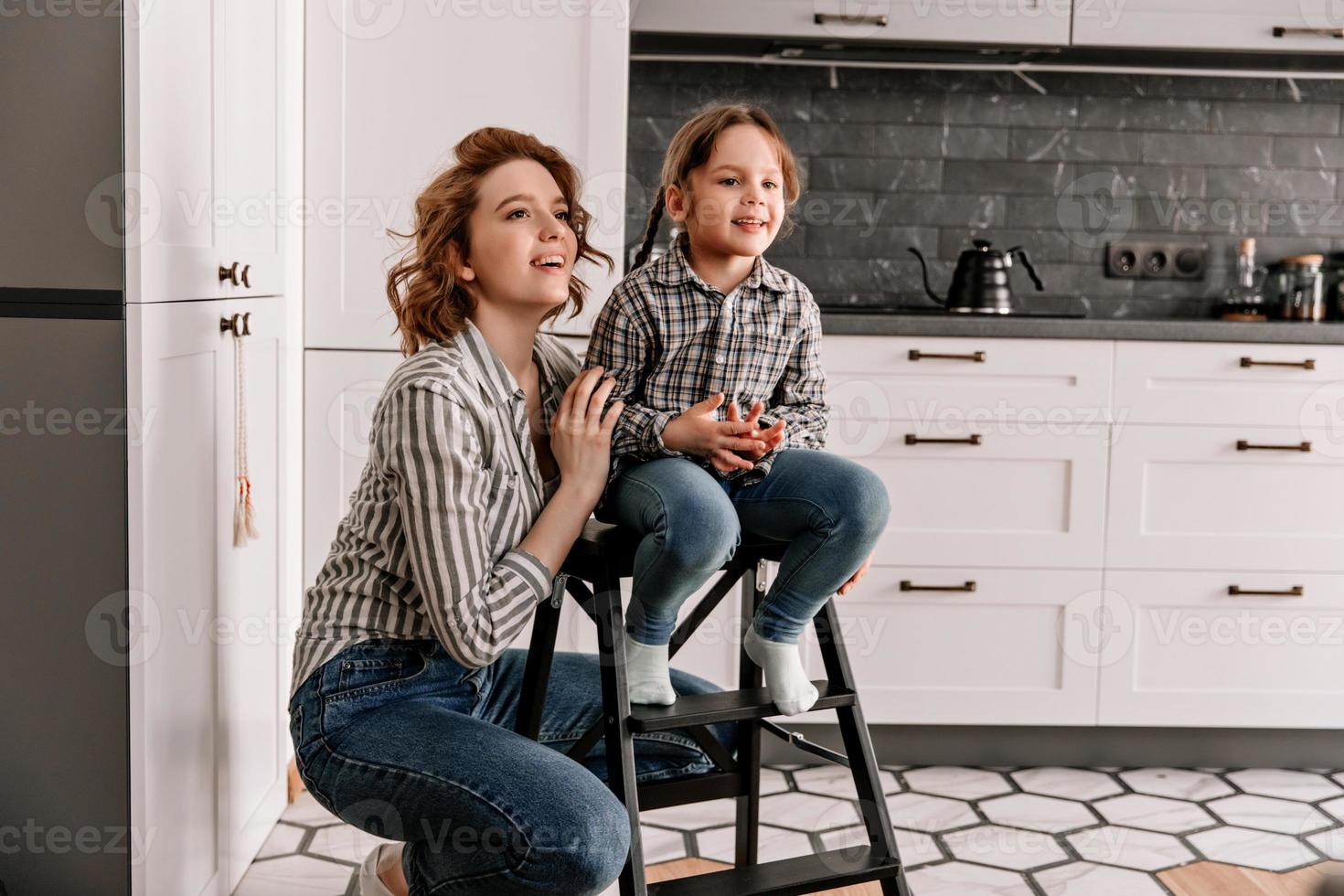 Girl sits on stairs and her mother poses next to against background of kitchen photo