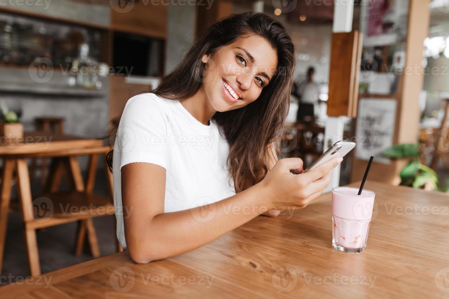 morena con teléfono en sus está descansando en el café. mujer con camiseta sonríe a ca 12923105 de stock en Vecteezy