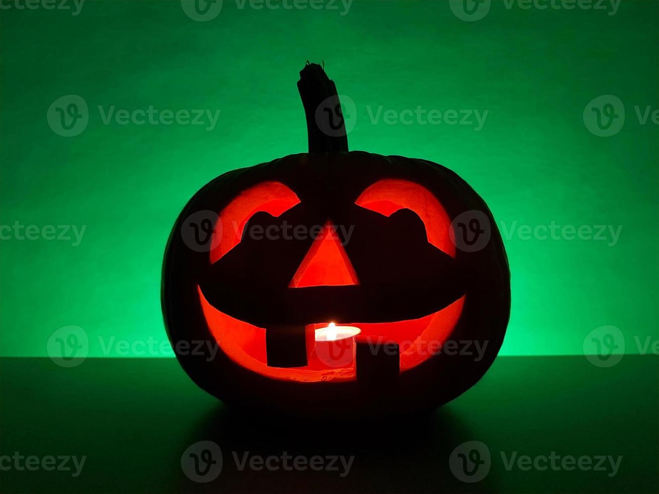 Halloween carved glowing pumpkin jack-o-lantern with candle inside stands on surface in dark night and blurred defocused background. The back wall highlighted in green. Spooky holiday concept. photo