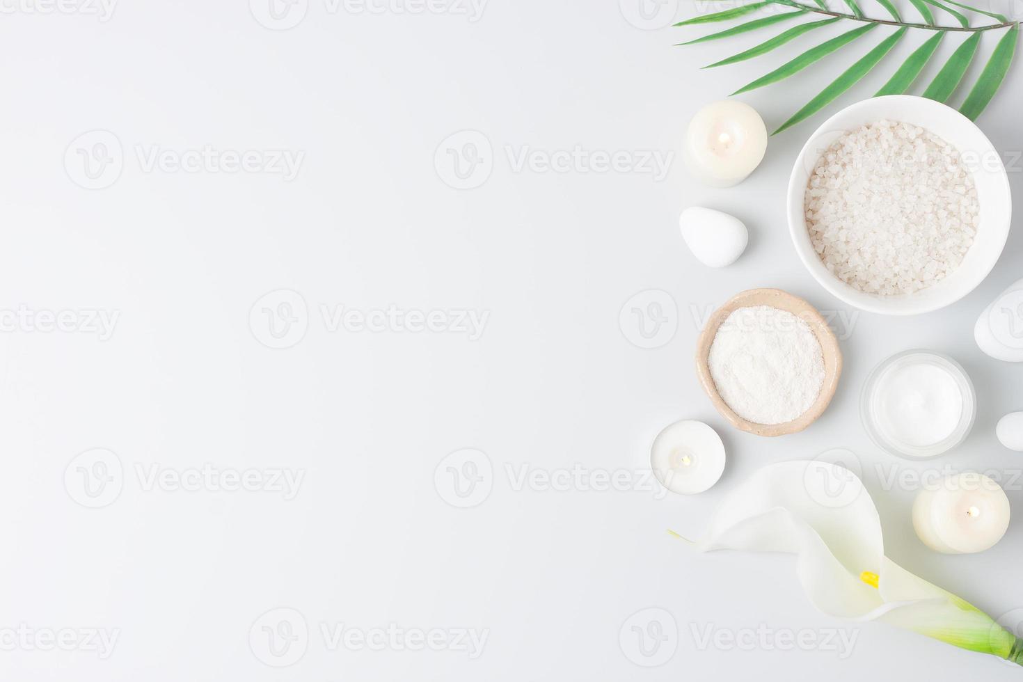 Spa background with bowl of salt, skin care products, white stones, candles, palm leaf and white flower on white. Flat lay, copy space photo