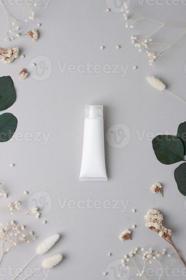 White tube of cosmetic cream with flowers and green leaves on grey background. Flat lay photo