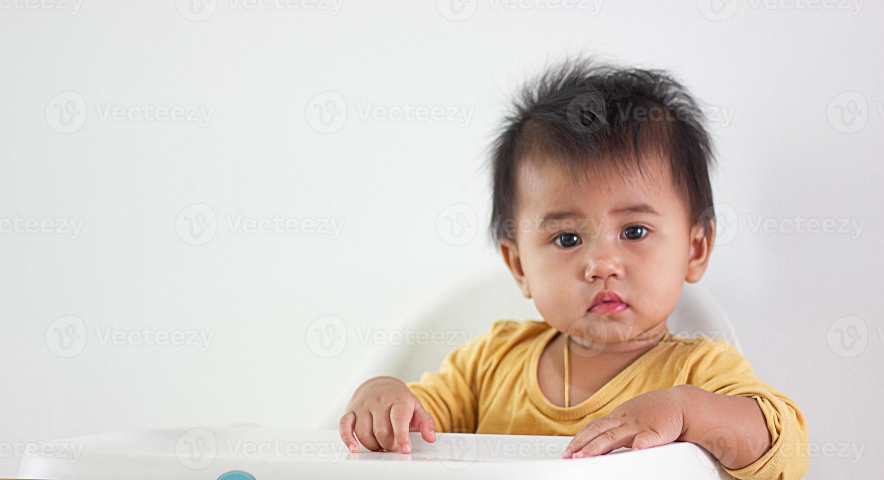 una niña de camisa amarilla de etnia asiática se sienta pacientemente en una silla sola en casa. expresión de la emoción, el entusiasmo, la infancia y la puerilidad. fuera esa linda guau cara felicidad vida foto