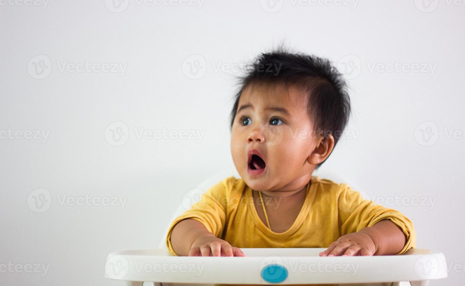 una niña de camisa amarilla de etnia asiática se sienta pacientemente en una silla sola en casa. expresión de la emoción, el entusiasmo, la infancia y la puerilidad. fuera esa linda guau cara felicidad vida foto