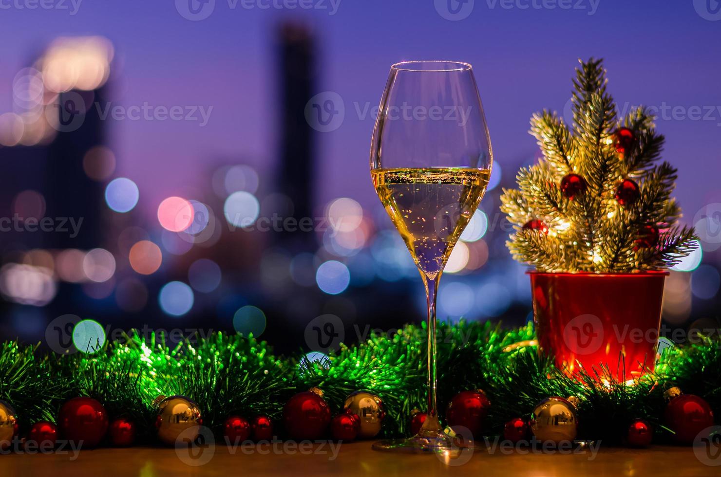 una copa de vino blanco se pone sobre una mesa de madera con un árbol de navidad decorado con adornos de adorno y luces sobre el fondo de las luces de la ciudad de colores. foto