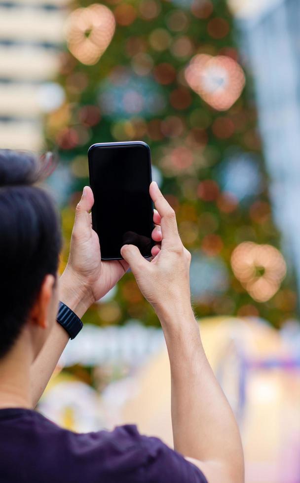 Asian man holding and taking Christmas tree photo with smartphone to enjoy Christmas holiday.