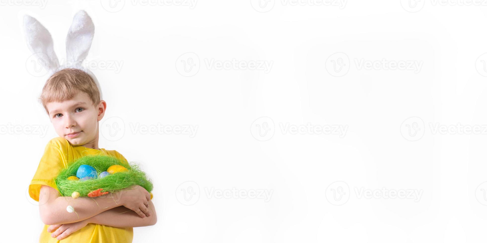 Happy Easter banner. Funny child boy in a white T-shirt in rabbit ears and with colorful Easter eggs on a white background with a place for text. photo