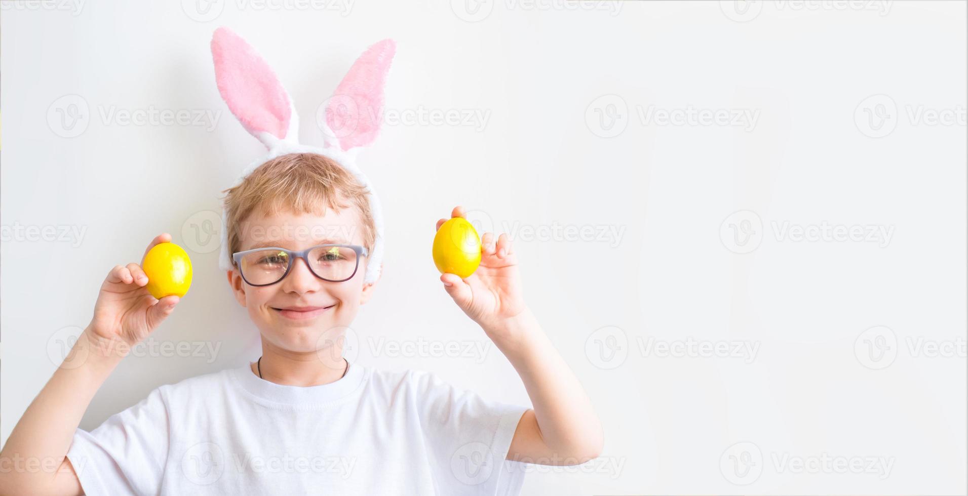 Happy child smiles in glasses in a white t-shirt in rabbit ears and with colorful yellow Easter eggs on a white background. Happy Easter concept. photo