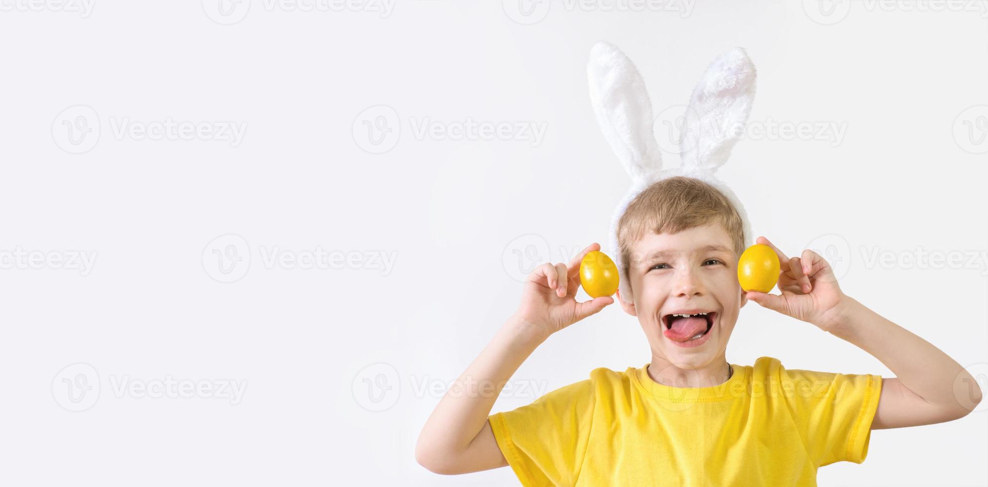 Happy Easter banner. Funny child boy in a white T-shirt in rabbit ears and with colorful Easter eggs on a white background with a place for text. photo