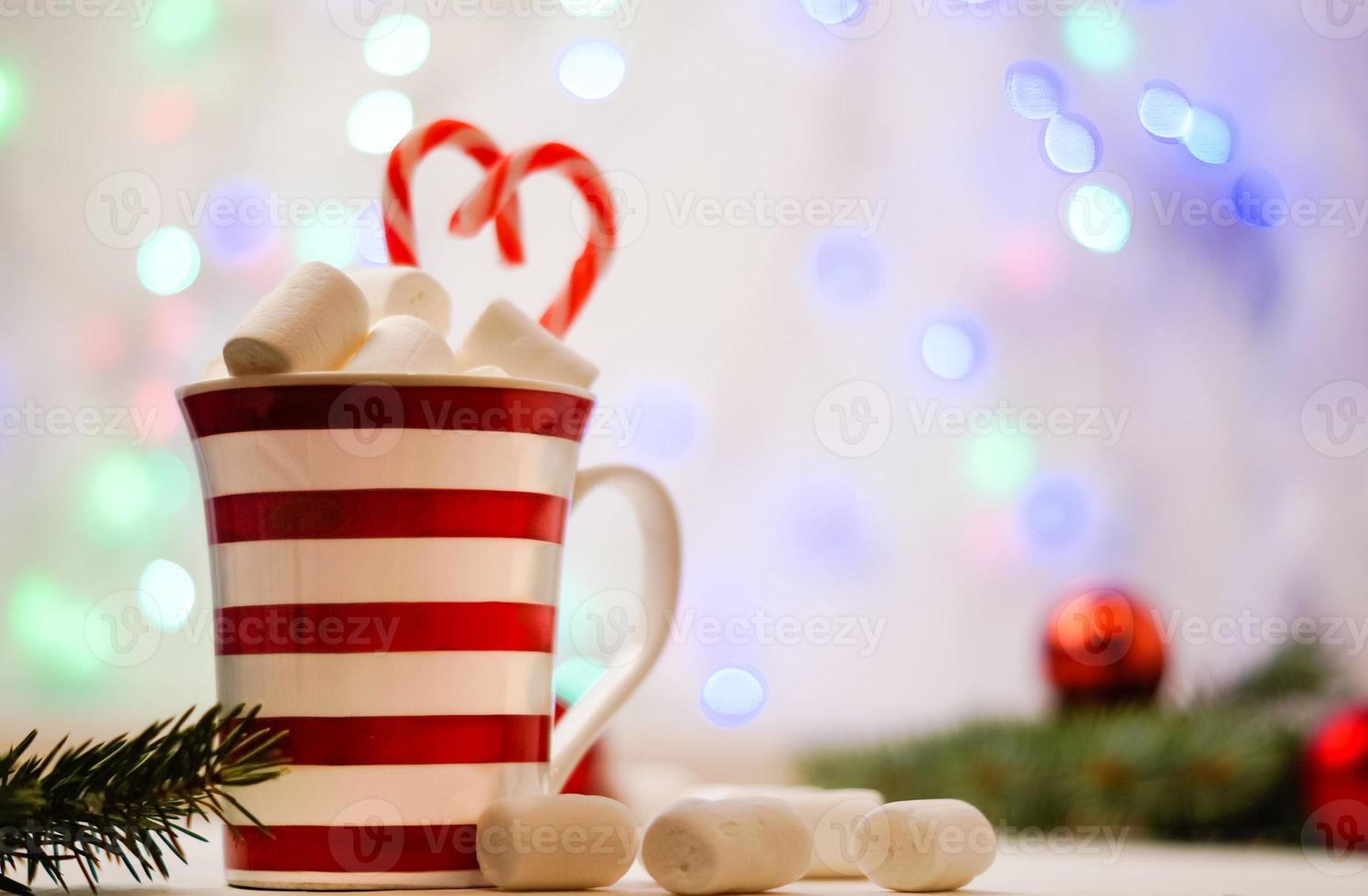 Hot cocoa with marshmallows and red-white lollipop. Hot chocolate with marshmallows in a New Year's cup against a background of multi-colored bokeh. photo