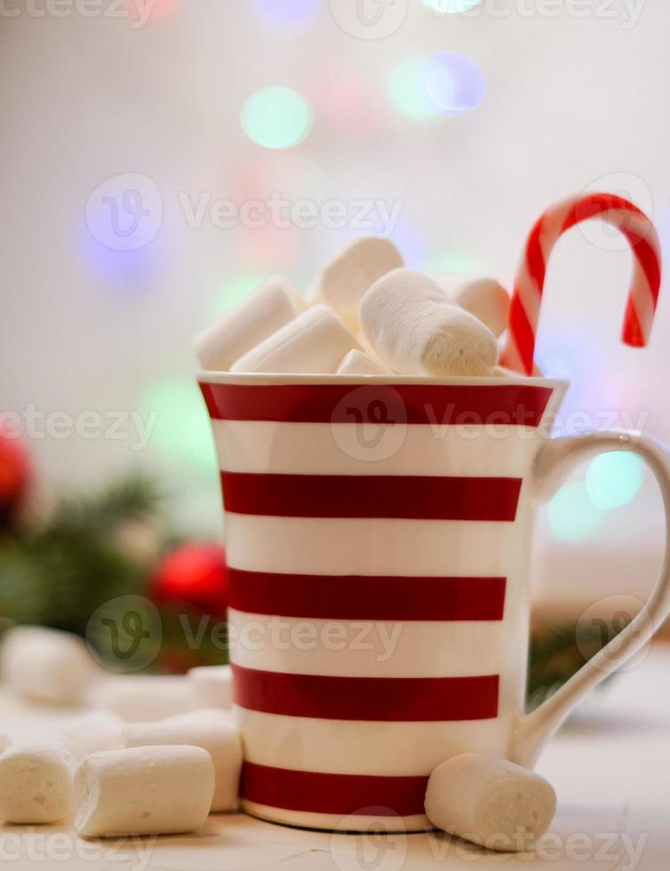 Hot cocoa with marshmallows and red-white lollipop. Hot chocolate with marshmallows in a New Year's cup against a background of multi-colored bokeh. photo