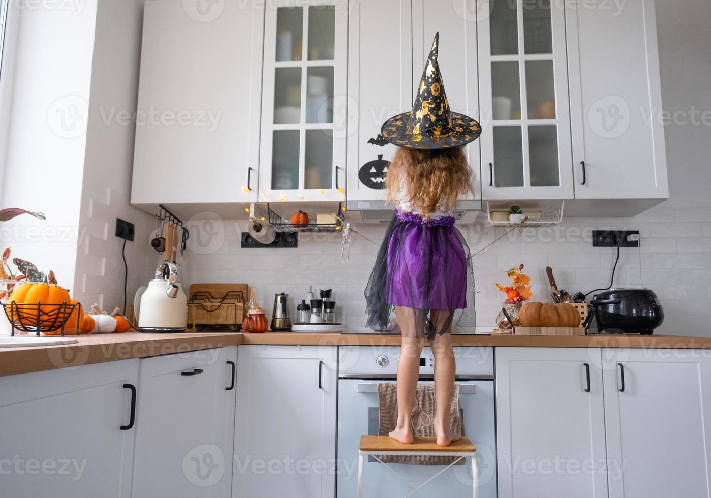 Child decorates the kitchen in home for Halloween. Girl in a witch costume plays with the decor for the holiday - bats, jack lantern, pumpkins. Autumn comfort in house, Scandi-style kitchen, loft photo