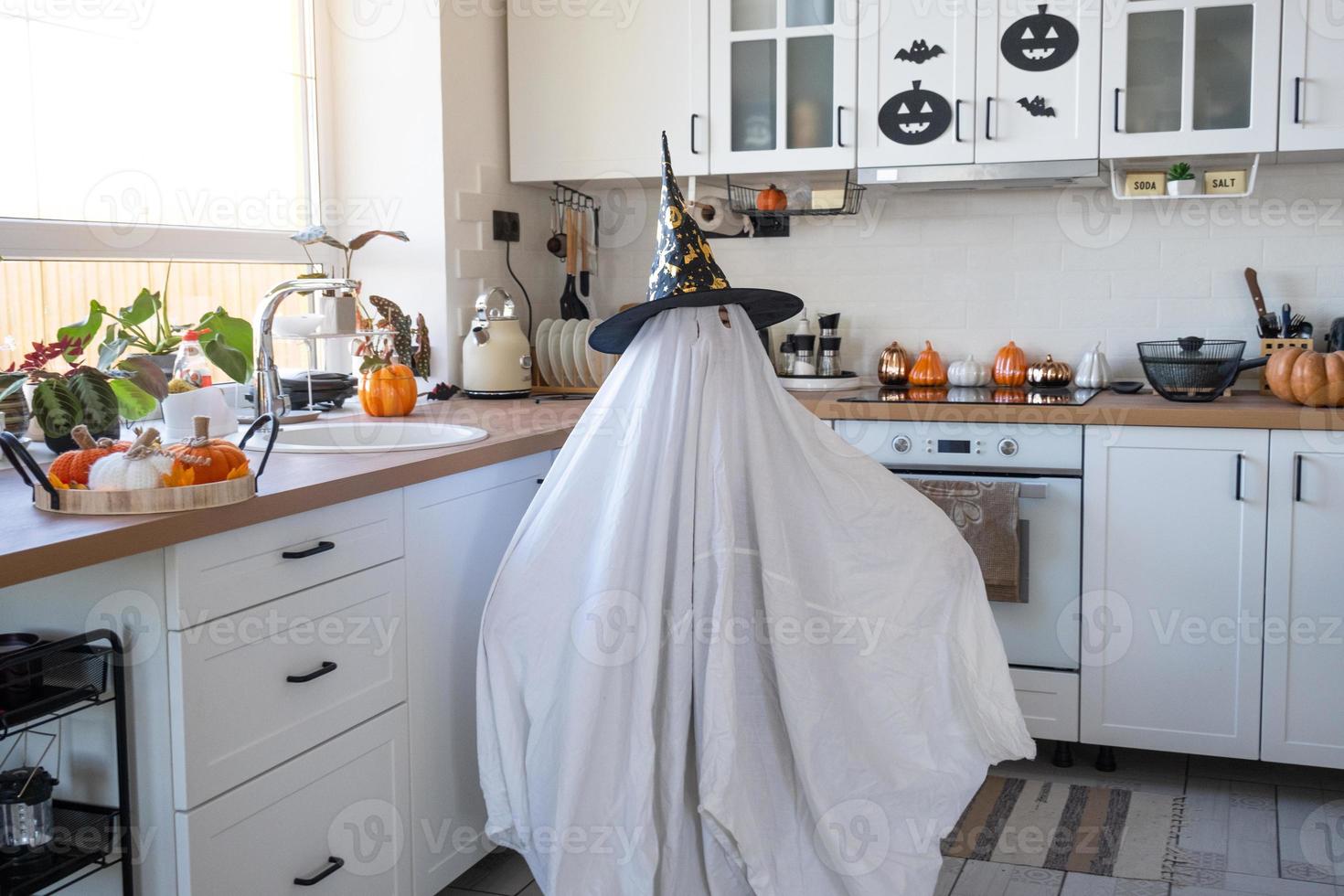 un niño en sábanas con ojos cortados como un disfraz de fantasma en la cocina decorada para la fiesta de halloween. un pequeño fantasma amable y divertido. fiesta de Halloween foto