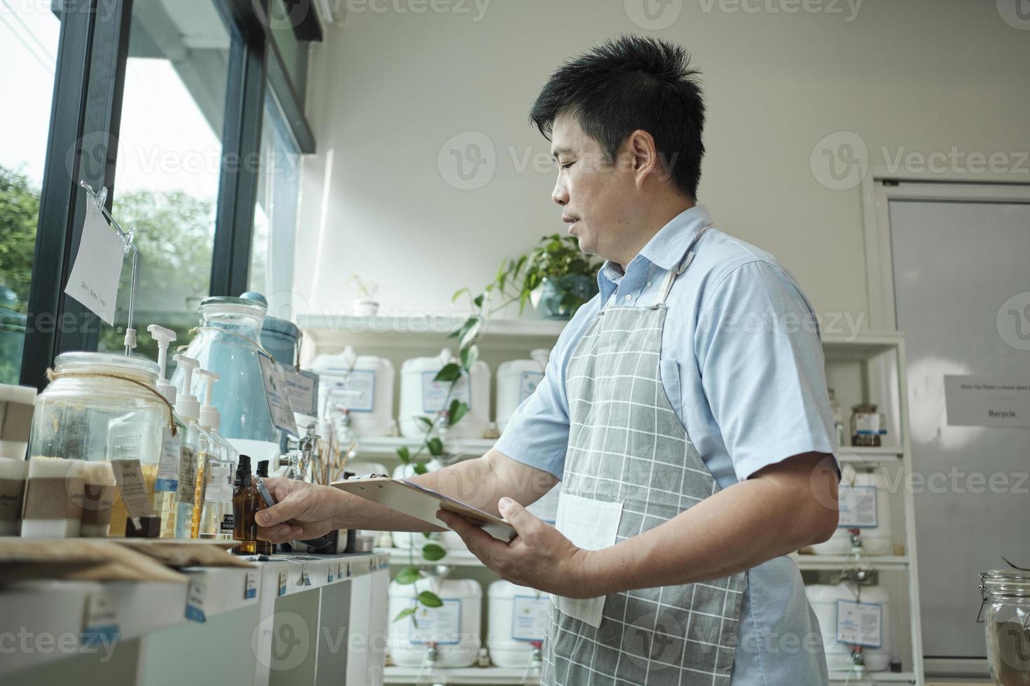 Asian male shopkeeper checks stock of natural organic products at window display in refill store, zero-waste and plastic-free grocery, eco environment-friendly, sustainable lifestyles, reusable shop. photo