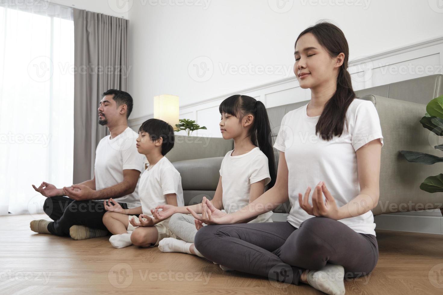 feliz familia tailandesa asiática, padres e hijos se sientan en el piso de la sala de estar, cierran los ojos juntos, meditan tranquilamente en posición de loto y practican yoga saludable, fin de semana de estilo de vida doméstico. foto