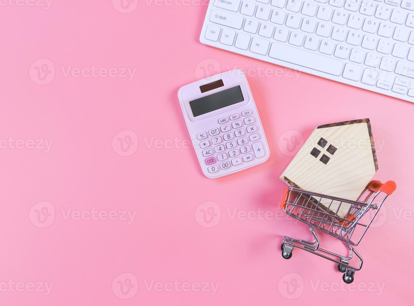 diseño plano del modelo de casa de madera en carrito de compras y calculadora rosa y teclado de computadora sobre fondo rosa con espacio de copia, concepto de compra de vivienda. foto