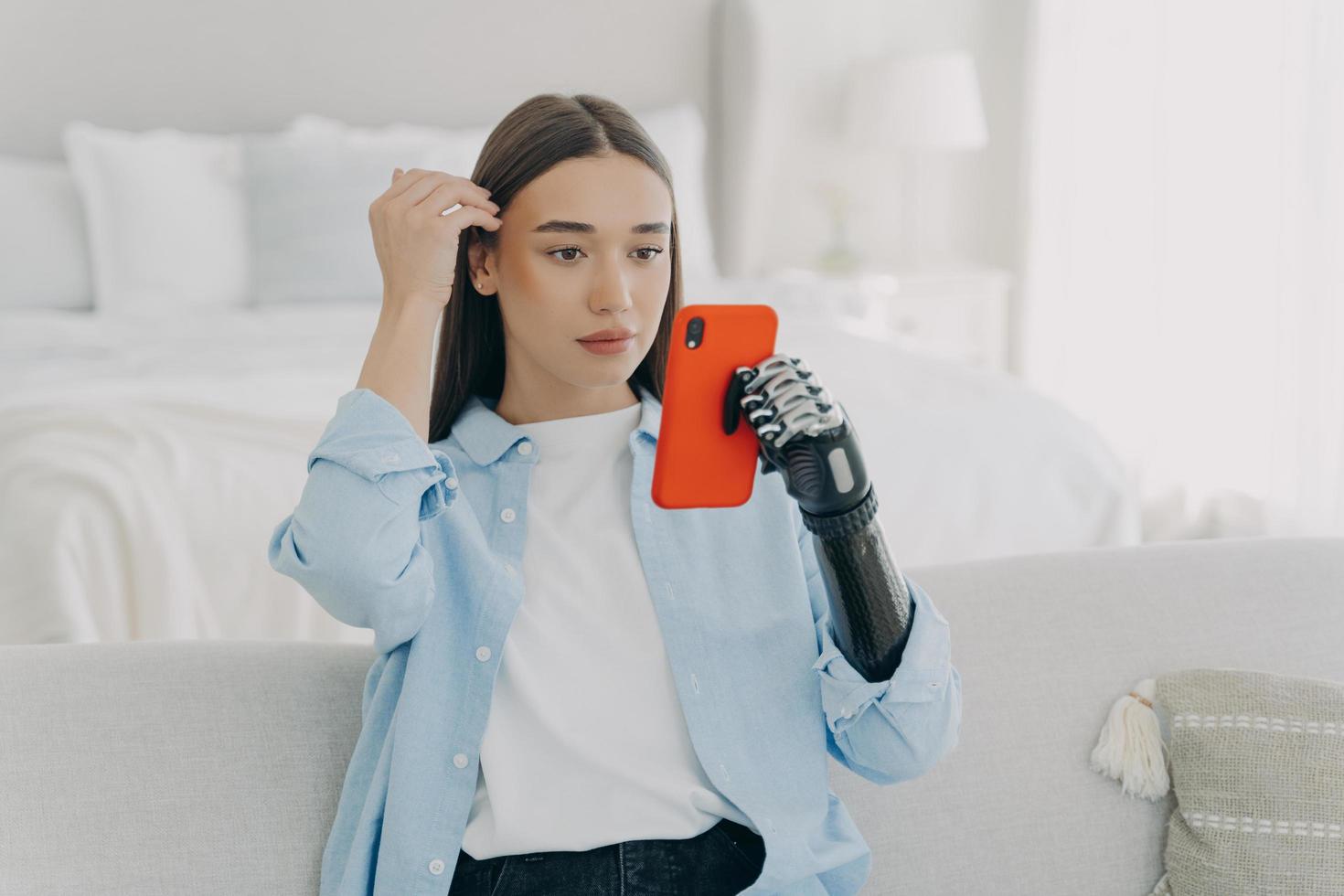 joven mujer discapacitada moderna sosteniendo un teléfono inteligente usando un brazo protésico biónico sentado en el sofá en casa foto