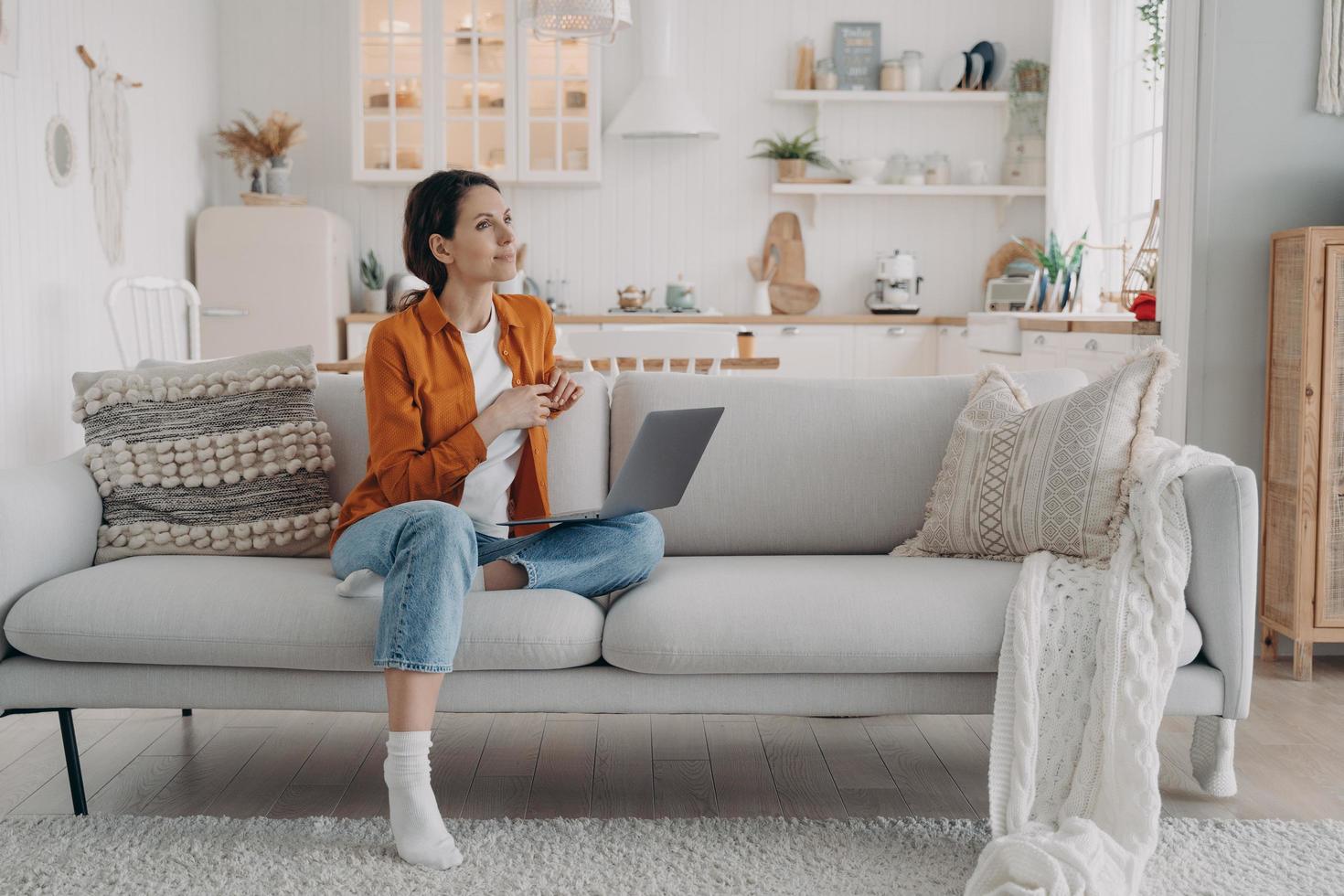 Pensive dreaming female rests with laptop on comfortable modern sofa in cozy living room at home photo