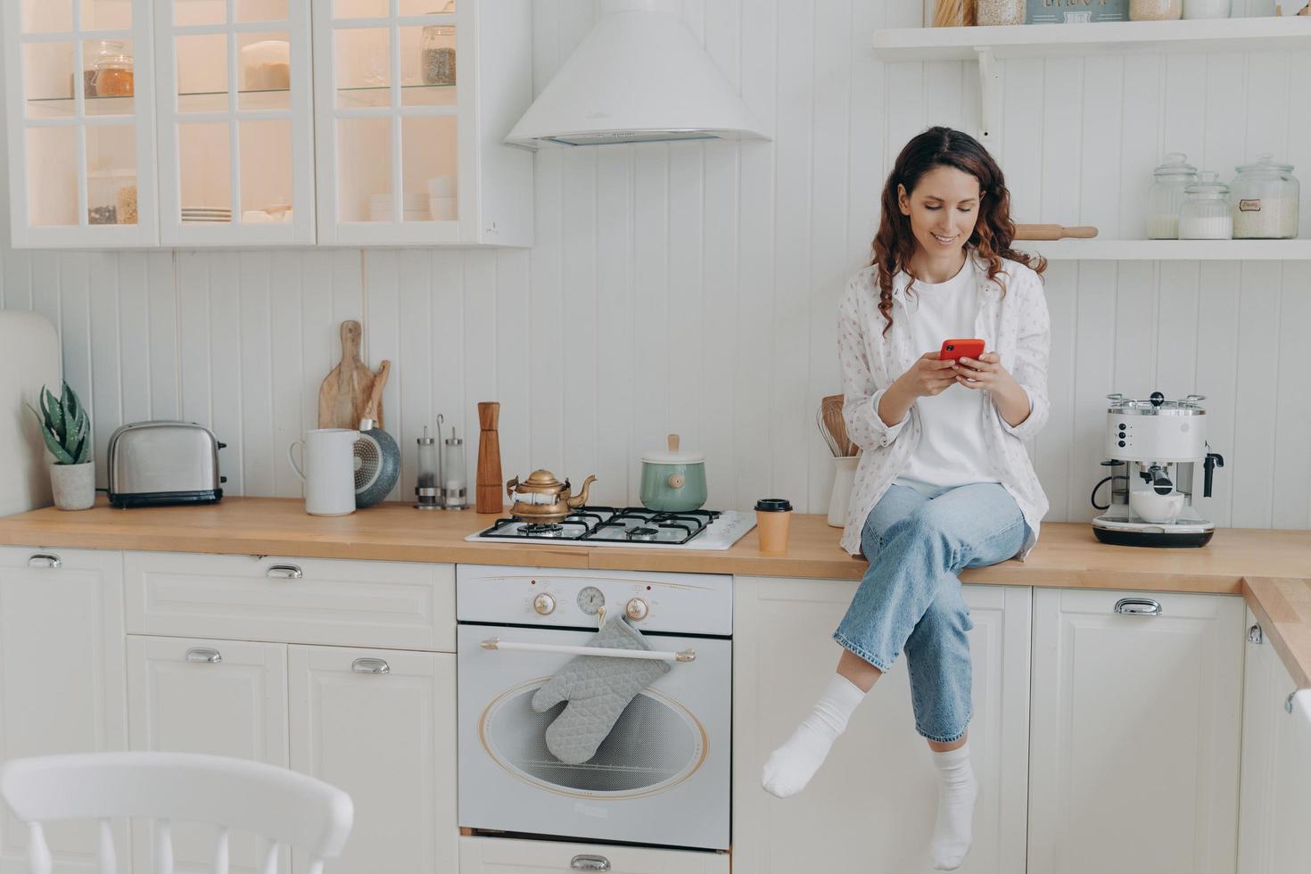 Female housewife satisfied with house cleaning services sits in modern clean kitchen with smartphone photo